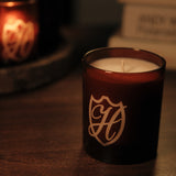 Unlit candle in glass vessel with H etched into the container, there is a lit version of the candle in the background