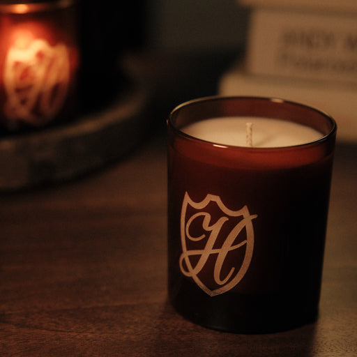 Unlit candle in glass vessel with H etched into the container, there is a lit version of the candle in the background