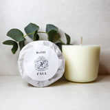 two wax candles one with paper packaging & a eucalyptus stem